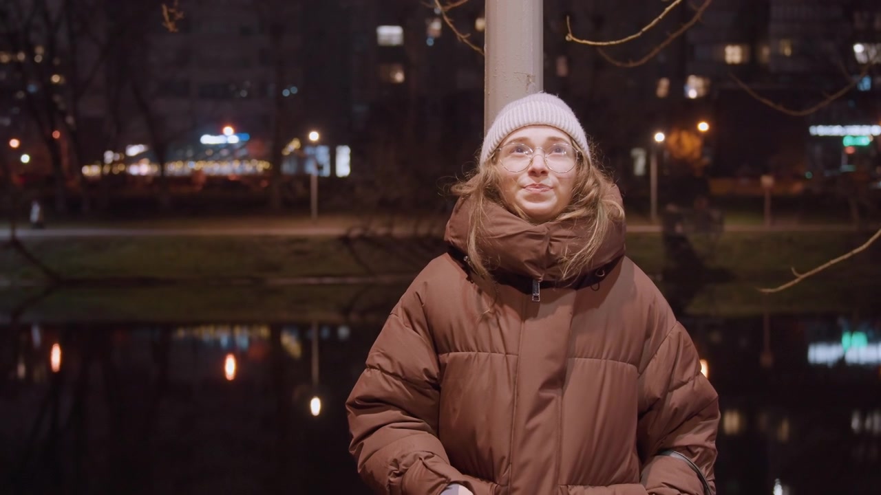 Белая женщина курит у озера ночью, задумчивая, в пуховике и шапке с очками, крупный план выдоха над отражениями в воде и городскими огнями, интимный портрет ночной мыслительницы.