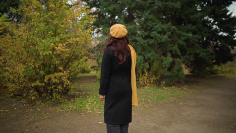 elegant woman in black coat with yellow beret and scarf stands in autumn park, looking around and touching her red hair with a warm smile