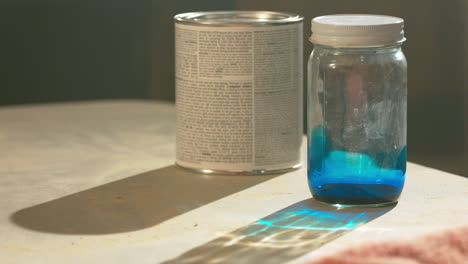 una mano colocando una lata de pintura y una botella de tinte azul sobre una mesa