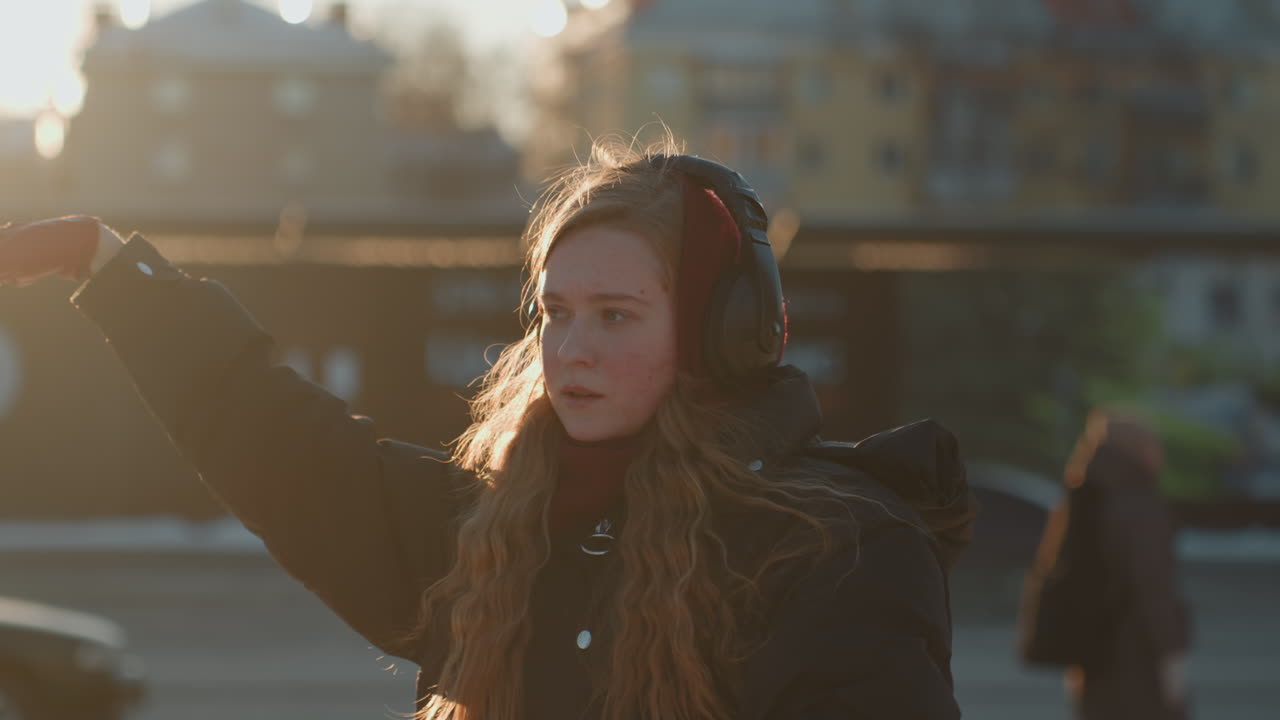 Artista al tramonto su una strada innevata al crepuscolo con cuffie e guanti rossi, gesti contemplativi e capelli retroilluminati che brillano nel sole ambrato, atmosfera urbana tranquilla e umore riflessivo.
