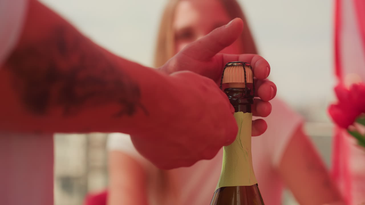 primo piano mani che stappano champagne donna caucasica, mani maschili tatuate rimuovono la gabbia metallica e afferrano la bottiglia, rilascio del tappo e accumulo di frizzante, donna sorridente sfocata sullo sfondo, bordo del tavolo rosso, tulipano