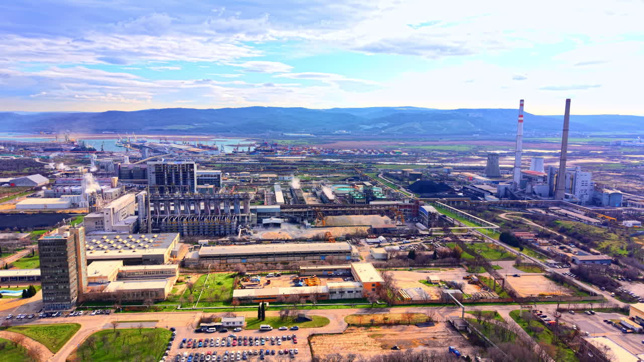 Amplia zona industrial con chimeneas humeantes, almacenes, estacionamientos y un puerto con grúas al fondo. Vista panorámica de una gran área portuaria industrial.