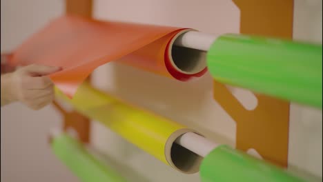 female hands pulling a vinyl material color orange from a wall stock of supplies of various rolls of paper