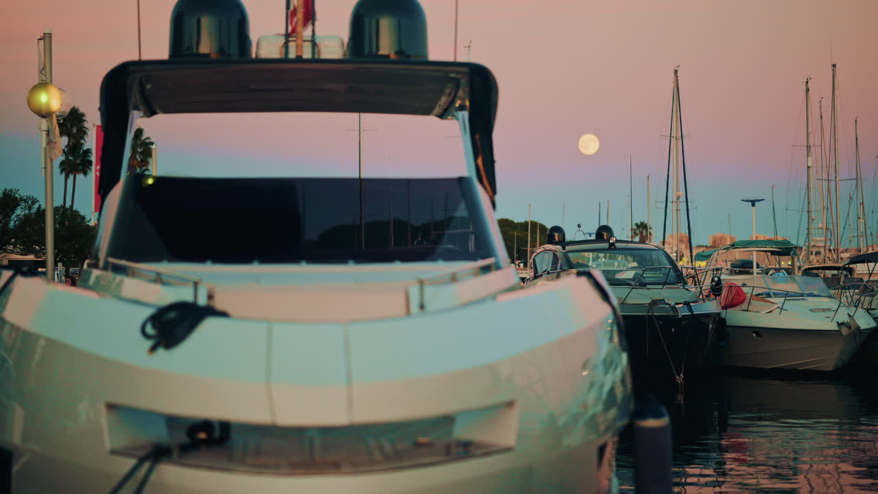 Front view of a sleek white yacht moored in the marina at twilight, with the full moon hanging in the soft sky behind it