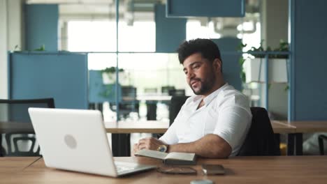 portrait-of-a-handsome,-stylish-young-man-of-Arab-descent-sitting-in-a-modern-business-center-office-with-a-laptop,-greeting-and-starting-a-video-call