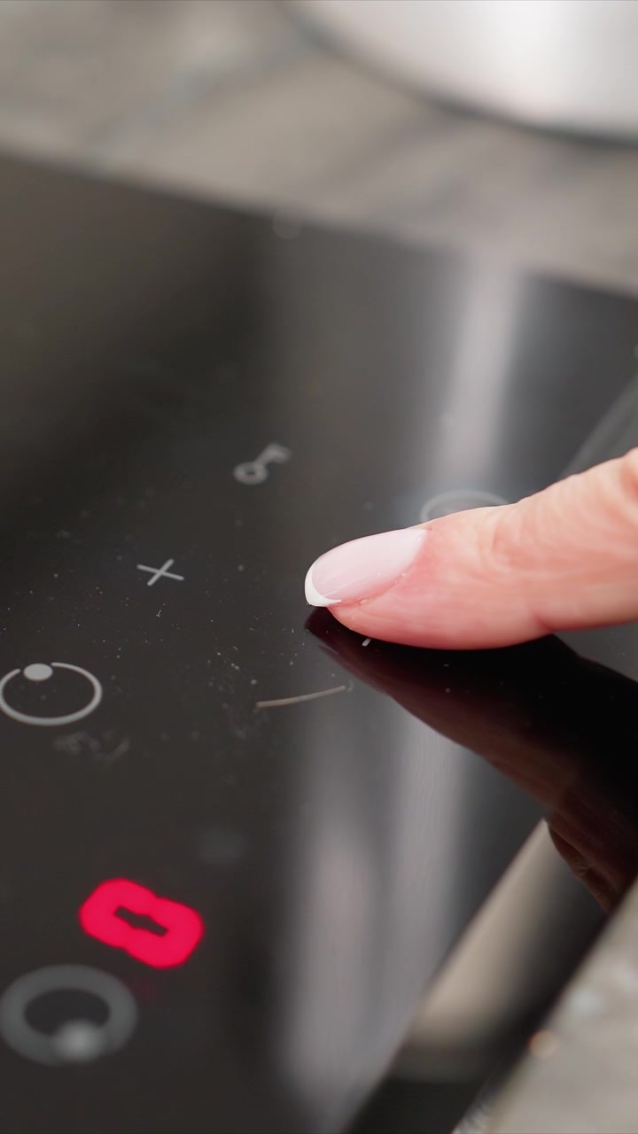 Primo piano di un piano cottura a induzione nero mentre le dita interagiscono con i controlli touch, attivando gli elementi riscaldanti e visualizzando indicatori rossi. La superficie liscia in vetro presenta simboli circolari.