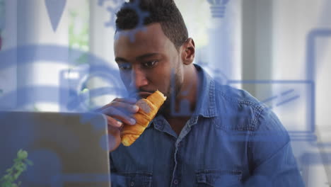 múltiples iconos digitales contra un hombre afroamericano tomando un bocadillo mientras usa una computadora portátil