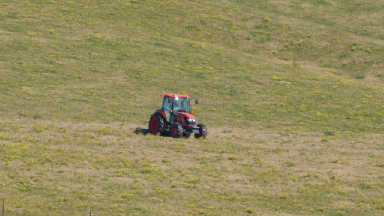 Red tractor moves steadily over sunlit grassy hillside, wide shot, natural daylight, rural Australia