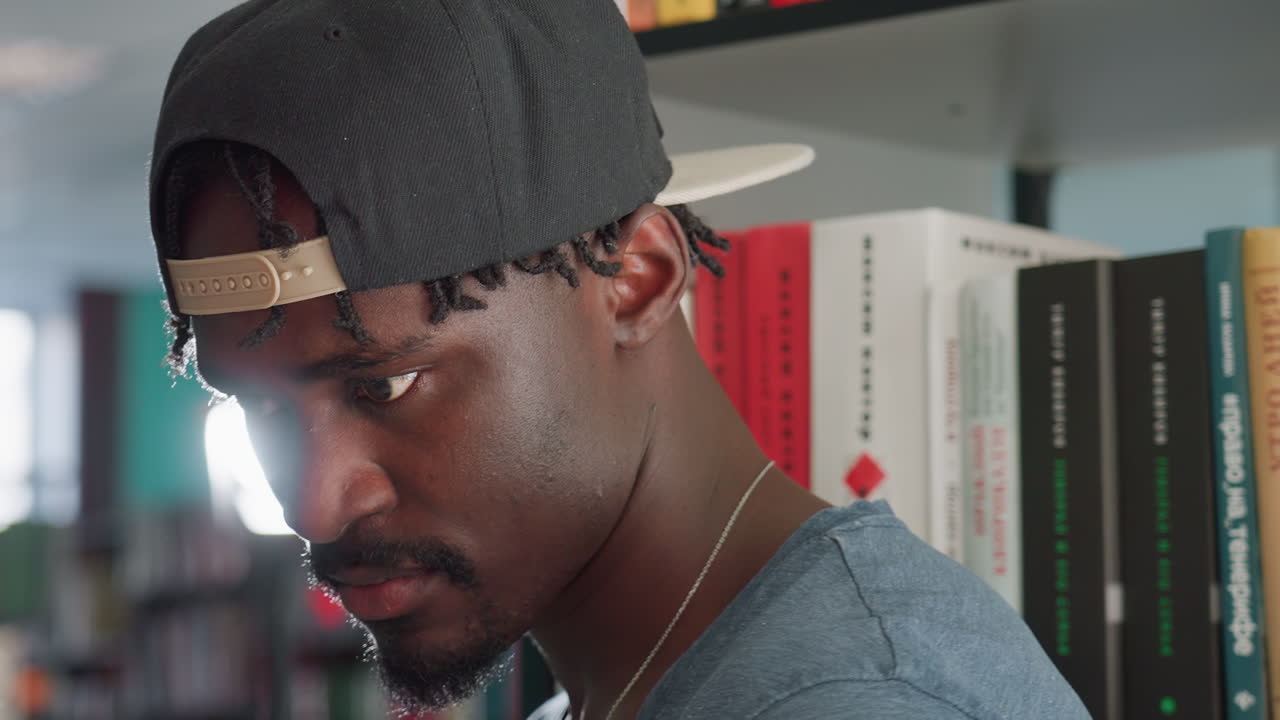 Close up side view of young man in casual clothing and backward cap standing near bookshelf, gazing downward with serious contemplative expression