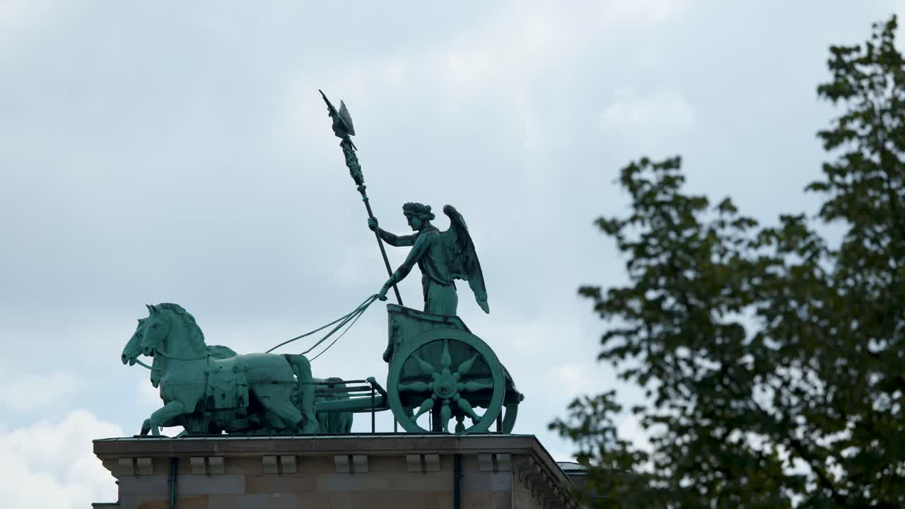 Static wide shot of quadriga statue atop Brandenburg Gate, partially obscured by leafy tree branches