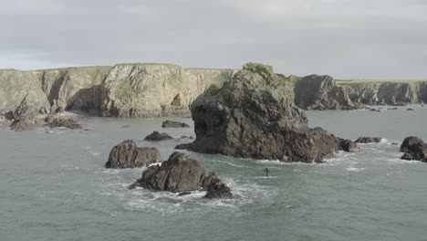 Trucking-aerial-tracks-paddle-boarder-in-ocean-chaos-near-jagged-rocks