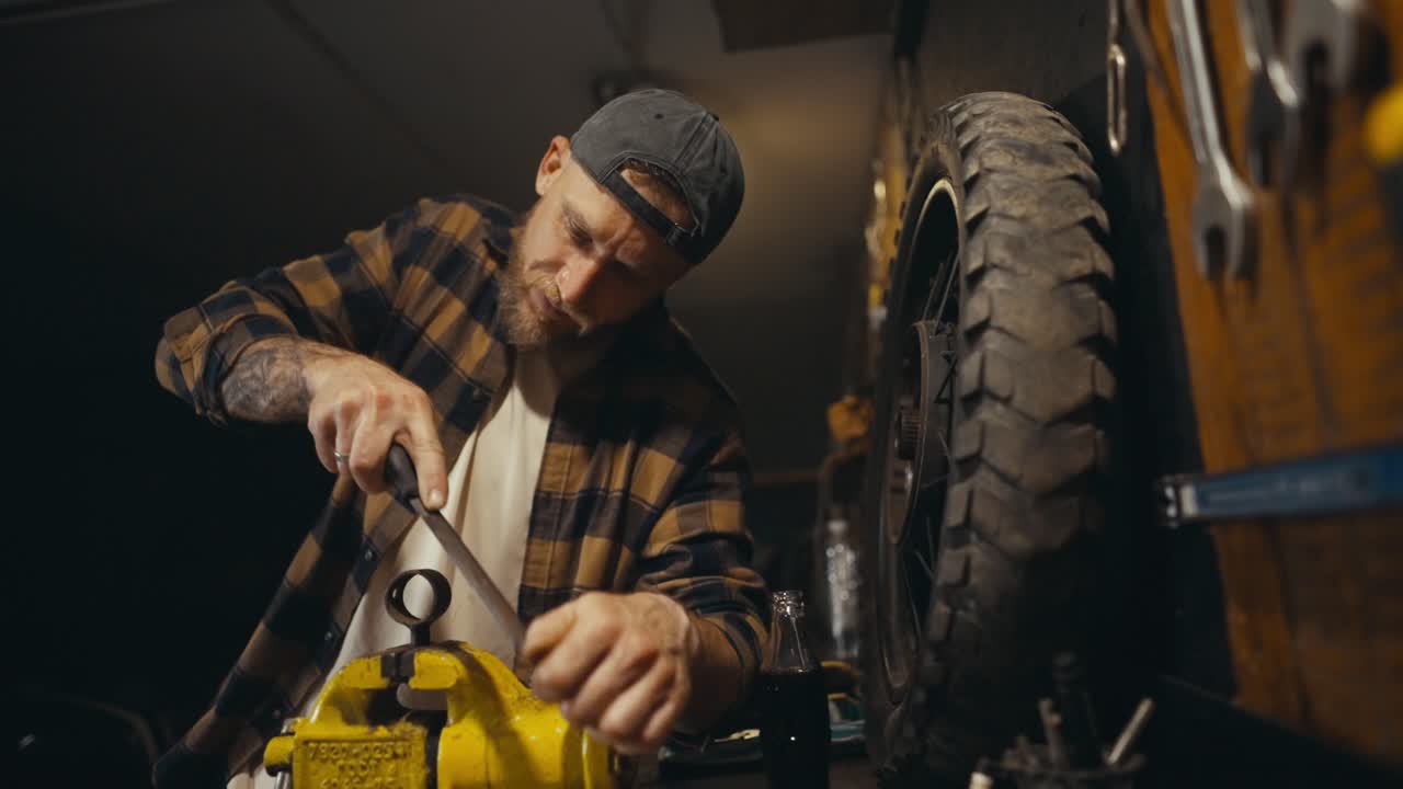A diligent guy mechanic with a beard works with a file on a workbench and drinks soda while repairing a part in his workshop