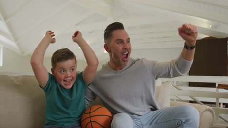 Happy-caucasian-father-with-son-watching-tv-and-holding-basketball-in-living-room
