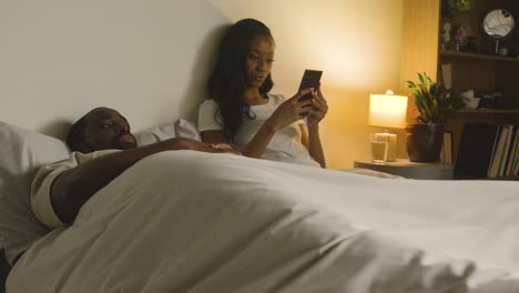 Pareja-En-Casa-Por-La-Noche-Acostada-En-La-Cama-Con-Una-Mujer-Mirando-El-Teléfono-Móvil