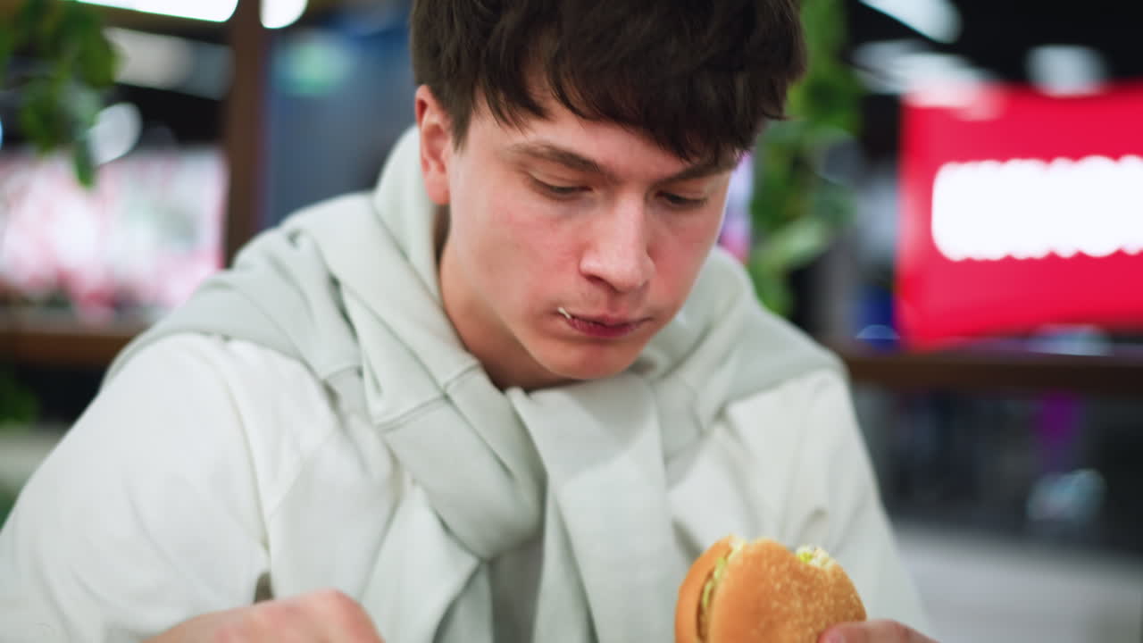 European man cleaning mouth with serviette after eating burger, closeup showing focused expression, indoor casual setting with soft background blur, hand movement wiping lips for hygiene