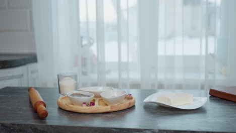 elegante mesa de cocina negra con bandeja de madera que contiene cuencos, especias, azúcar y mantequilla, rodillo colocado al lado del libro de portada marrón, cortina transparente blanca revela la vista del edificio a través de la ventana