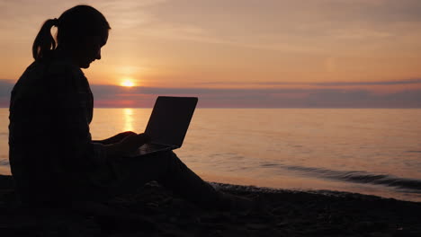 Silhouette-Einer-Frau,-Die-Bei-Sonnenuntergang-Mit-Einem-Laptop-Am-Meer-Arbeitet