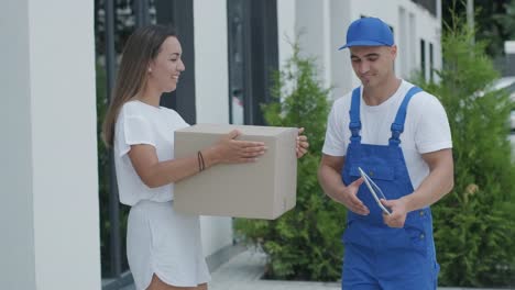 beautiful woman meets a delivery man who gives her a parcel box beside her home, she signs electronic signature on tablet. concept of courier, home delivery, online shopping