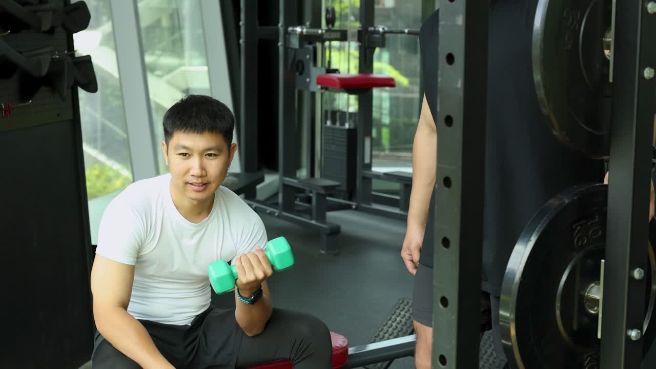 A man exercises with dumbbells in a well-lit gym, interacting with a trainer. The setting is modern and energetic