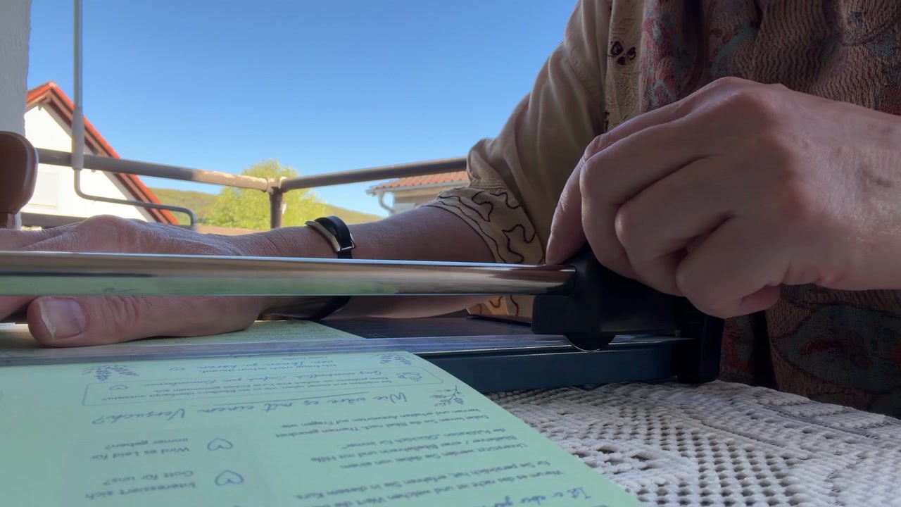 Close-up of a woman sitting on a balcony under a blue sky, carefully positioning and measuring paper in a paper cutter before starting to cut. Static shot.