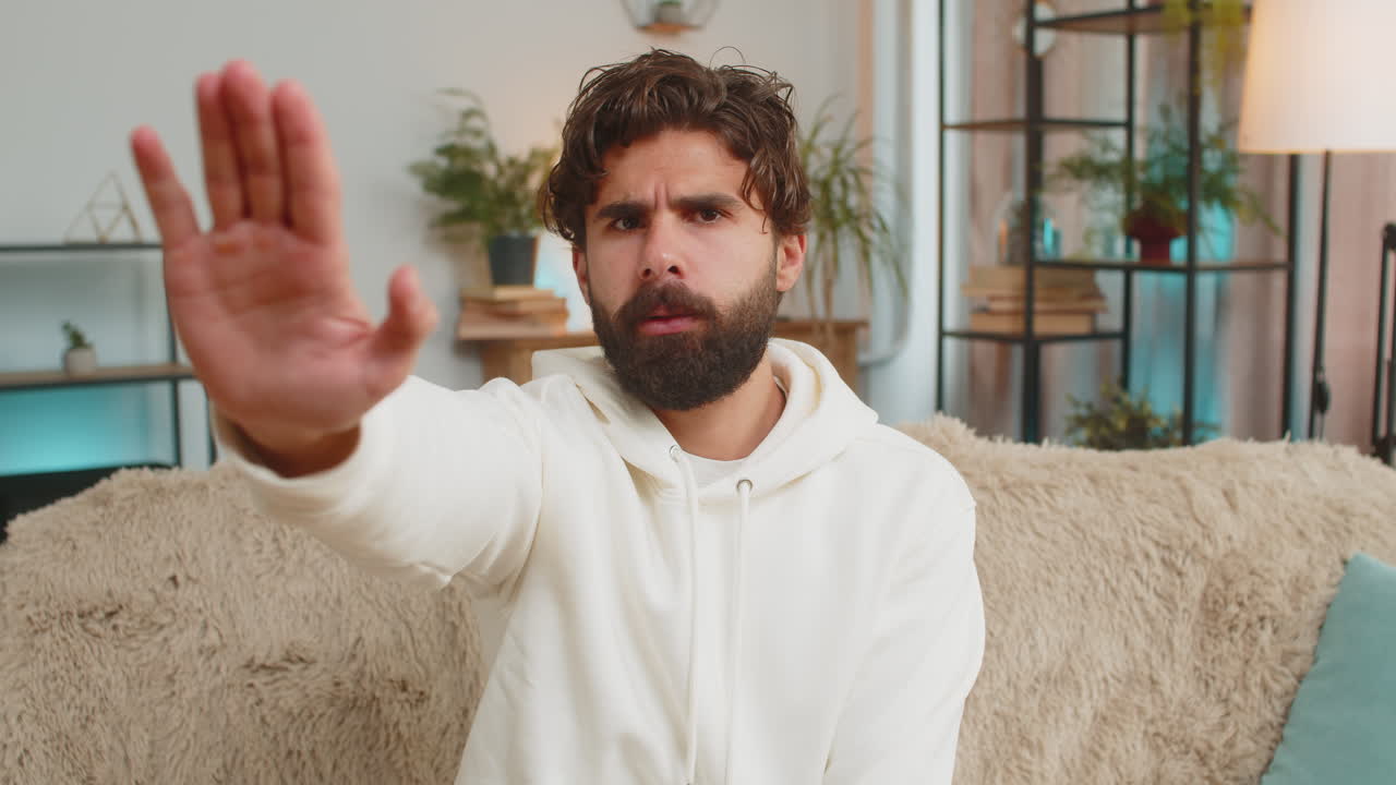 Indian young man guy on sofa couch with serious expression doing stop gesture with hand at home