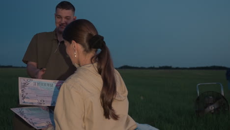 pilot extending hand to young man while handing certificate on grassy field at twilight, woman clapping beside deflated balloon fan in background celebrating achievement