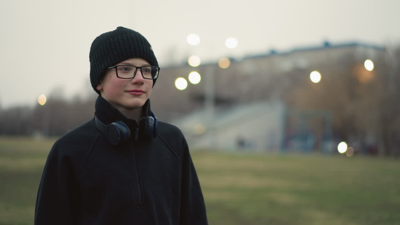 A coach holding a soccer ball in a field with a whistle in his mouth, playfully throws the ball at a young boy wearing glasses and a beanie in a stadium, then collects it back