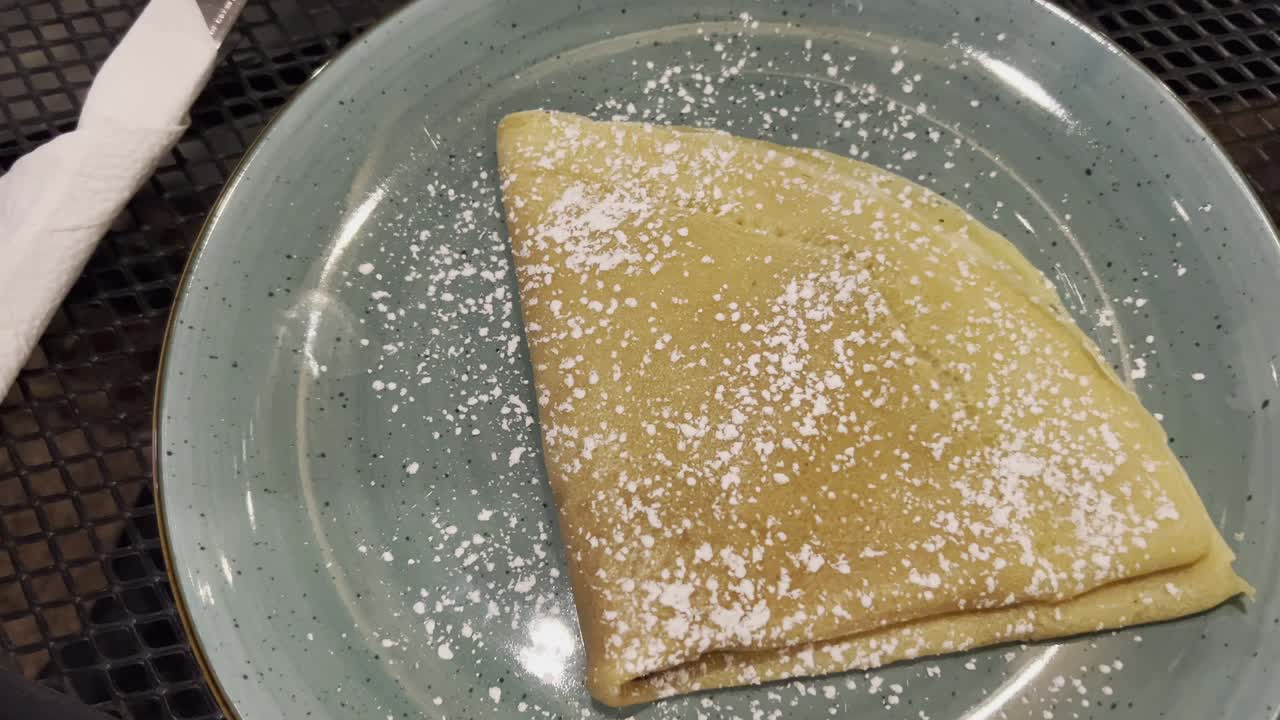 은 크레프, 부드럽게 설탕 가루를 뿌리며, 세련된, 얼룩진 밝은 푸른색 접시 위에 누워, 쾌락을 초대하고 맛있는 요리 경험을 약속합니다.