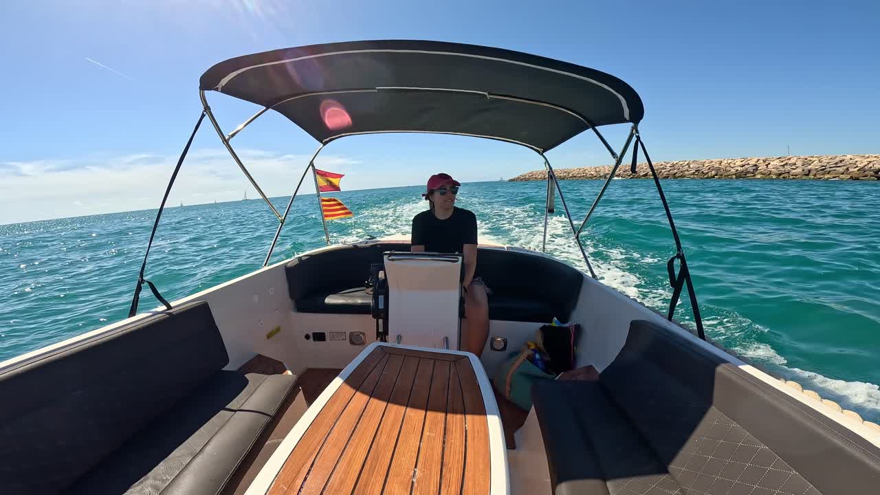Young woman drives a recreational boat on a sunny day, ocean and breakwater behind. A Spanish flag flutters at the rear under a clear blue sky