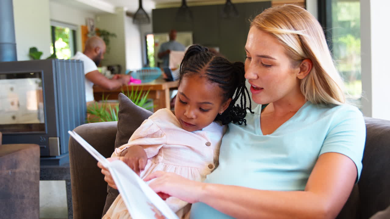 Pregnant Mother And Daughter Reading Book At Home Together With Family In Background