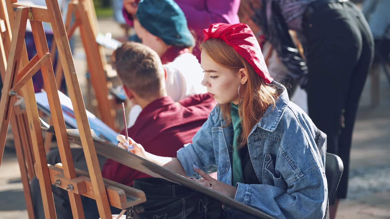 Young artists paint on easels in the city. Pretty woman artist is painting picture with brush and watercolors in the busy street. Side view of female artist.