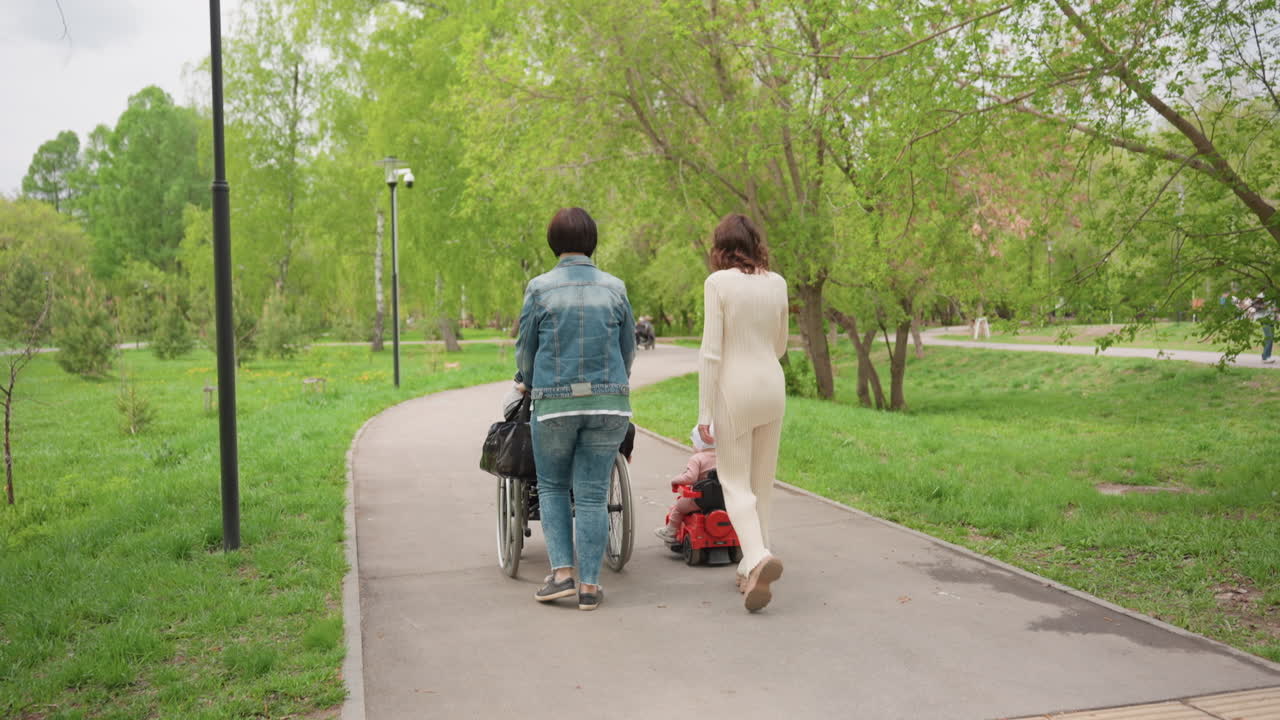 Park Path Women Walking With Wheelchair And Toddler On Scooter, TreeLined Walkway, Lamp Posts, Green Spring Foliage, Gentle Companionship, Supportive Interaction, Casual Clothes, Inclusive Outing