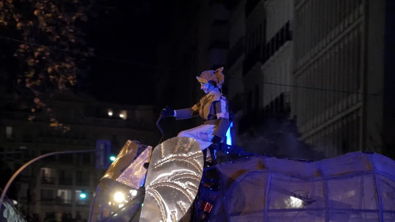 A man in a traditional jester-like costume, wearing a mask, rides a horse-like float in the Cabalgata de Reyes (Epiphany Parade). The clip begins blurred and pans as he looks left to right.