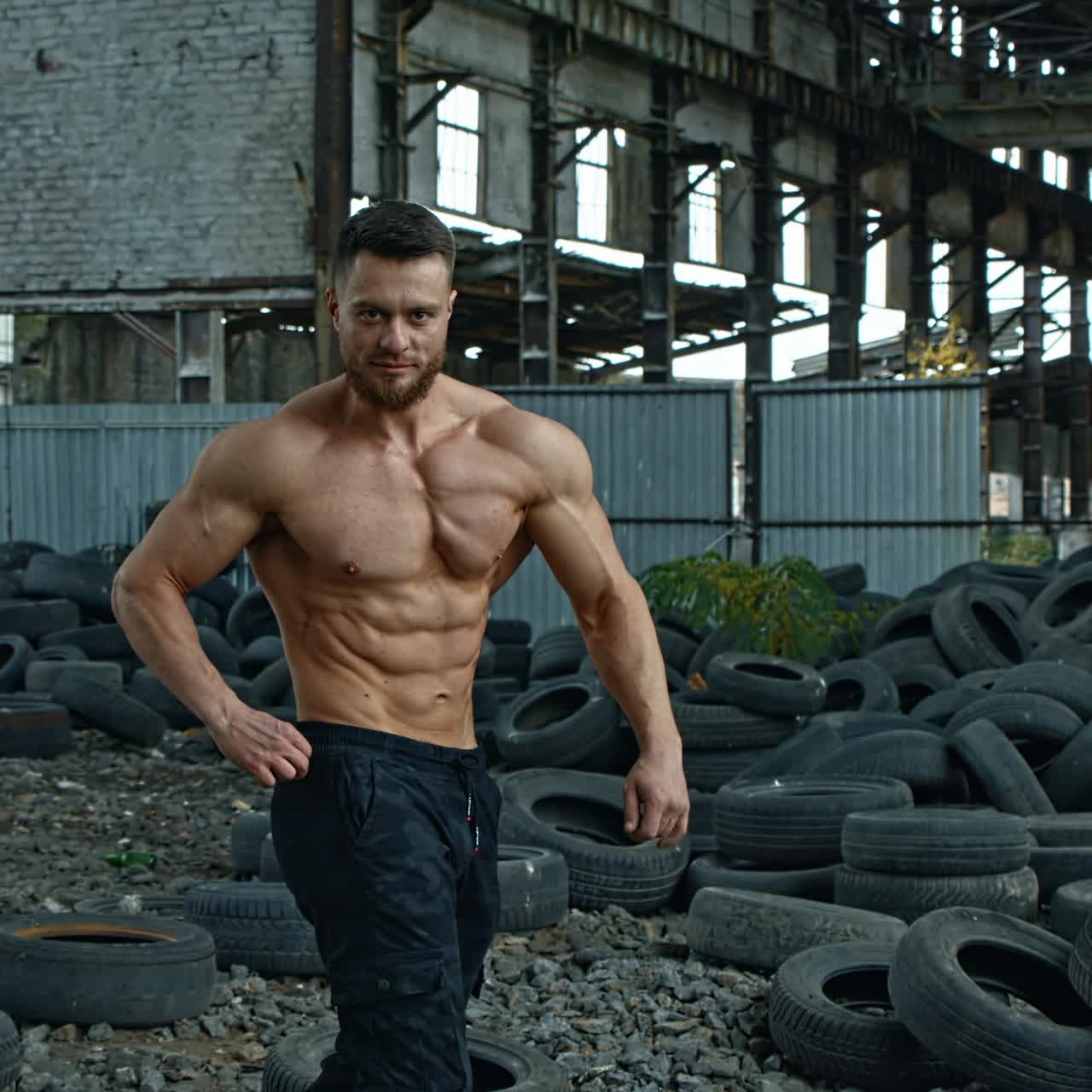 Shirtless bodybuilder in abandoned place. Young athlete with trained muscles posing to camera on used car tires background.