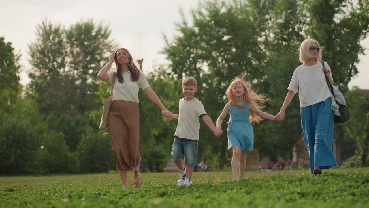 women with little ones holding hands jumping happily across green park field, smiling mothers and kids enjoying outdoor family time, casual summer clothes, tree background, playful movement