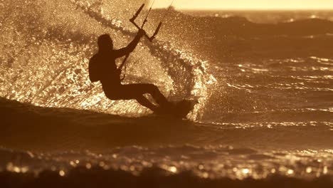 medium shot of a silhouetted person kitesurfing in the ocean with a lot of waves and kicks up a giant splash of water that is lit by the sun