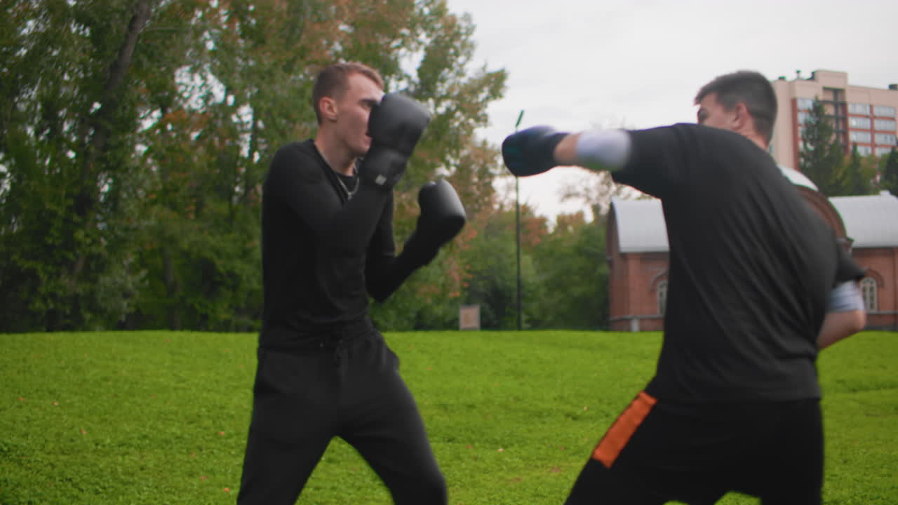 To hvide mænd, der træner med handsker, energisk udendørs boksetræning på en park, træneren holder fokus pads, mens fighteren slår jabs, hooks og uppercuts, defensive slips og kraftfulde.