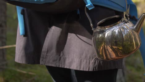 Metal-shiny-tea-pot-with-reflection-of-the-forest-and-autumn-slow-motion-atmosphere,-blue-tourist-bag,-through-trees