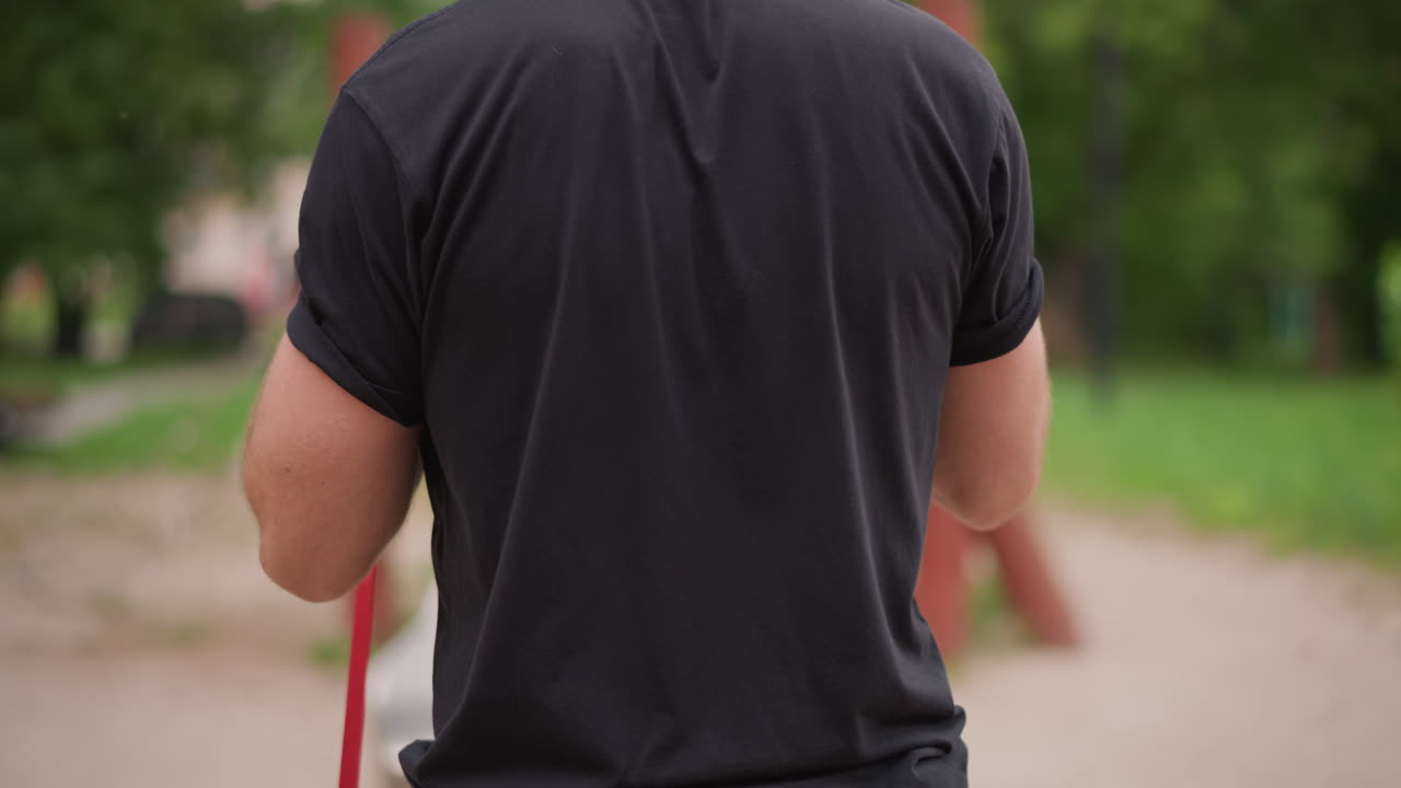 White Man Coach Adjusting Red Resistance Band Behind Back, Casual Park Scene, Methodical Looping And Anchoring, Relaxed Warmup Before Workout, Green Trees And Pathway In Background