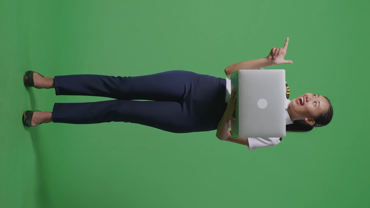 cuerpo lleno de piloto mujer asiática usando una computadora portátil y apuntando a un lado mientras está de pie en el estudio de fondo de la pantalla verde