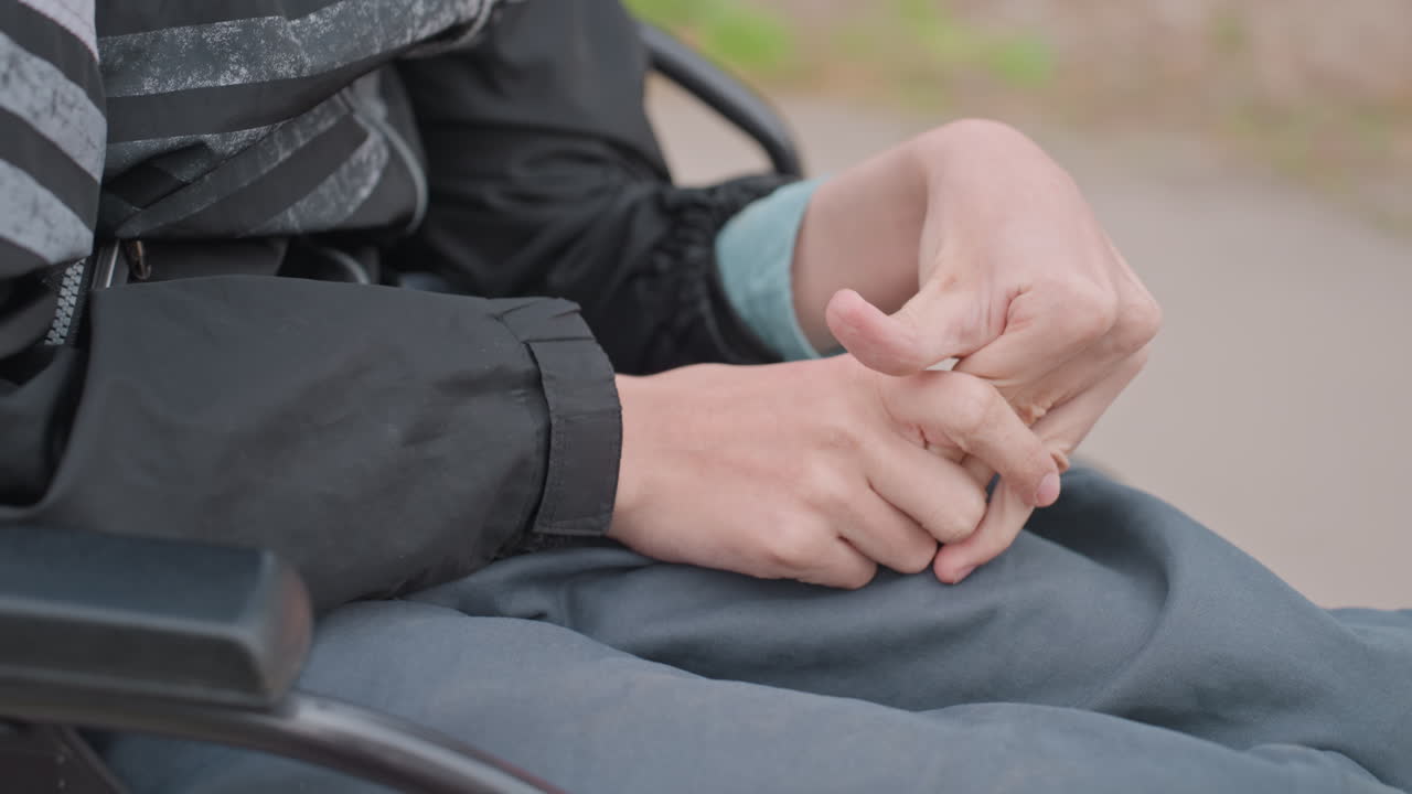 CloseUp Hands Twisting Thumb On Lap In Wheelchair, Subtle Nervous Motion And SelfSoothing Behavior, Casual Jacket Sleeve Visible, Neutral Park Background Conveying Intimate Emotional Detail