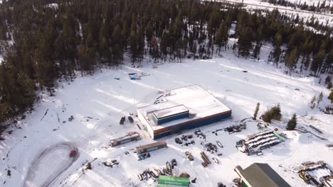 aerial view of a construction site with a blue warehouse building in the center surrounded by snow and dense forest