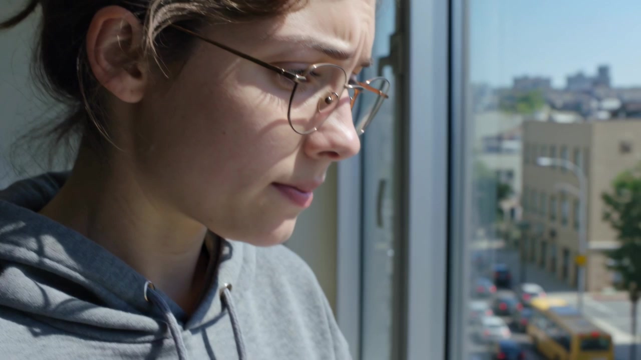 La telecamera si allontana, una donna con cappuccio grigio e occhiali preme le labbra contro la finestra, guardando autobus gialli.