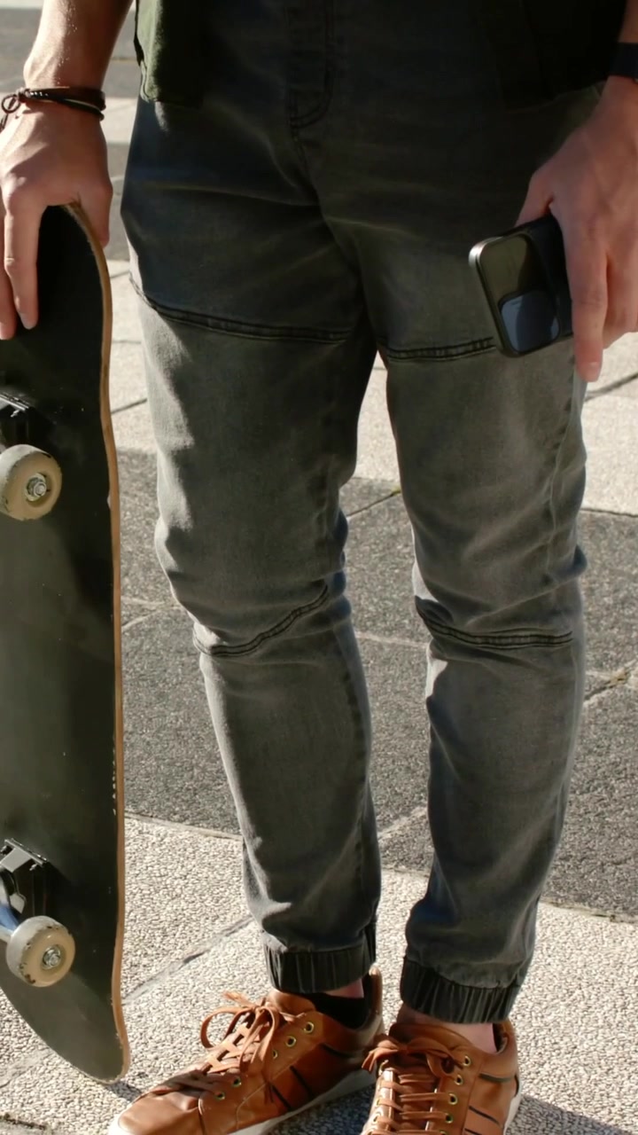 Vertical video: Adjusting man lowering skateboard on urban plaza, preparing to skate, holding phone