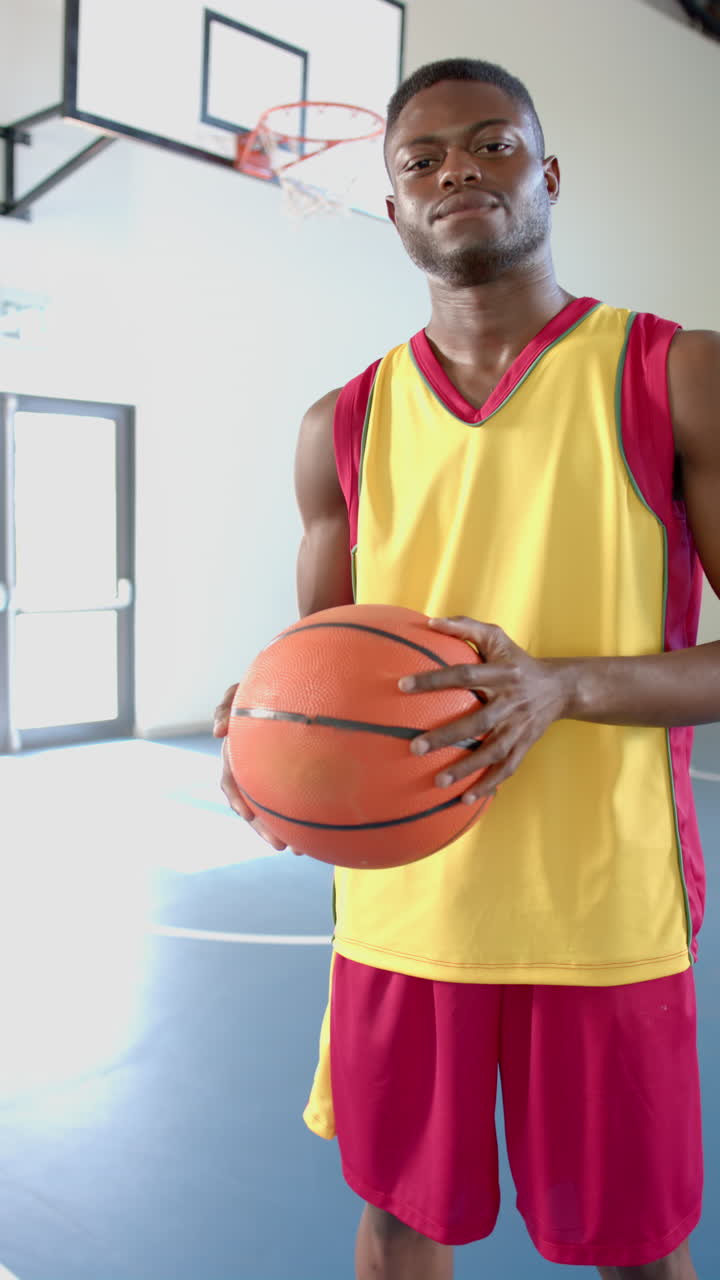 Vídeo vertical: Atleta masculino segurando uma bola de basquete, em pé em quadra coberta, de uniforme.