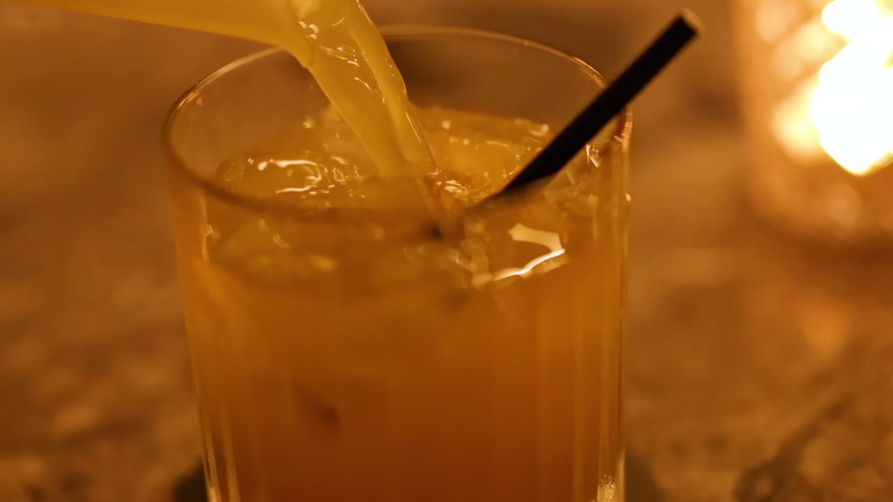 A drink is poured into a glass with ice and a straw, set against a warm, glowing background.