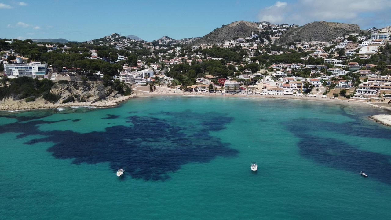 La discesa di un drone aereo rivela una vista a volo d'uccello di barche di lusso ancorate nelle acque turchesi della spiaggia di El Portet a Moraira, Costa Blanca, Spagna.