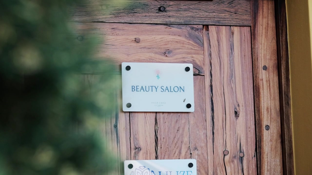 Letrero de salón de belleza en una puerta de madera rústica que se revela detrás de follaje verde.
