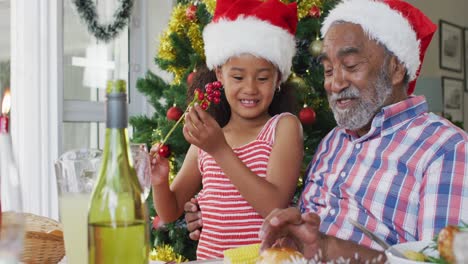 Happy-african-american-grandfather-and-granddaughter-wearing-santa-hats-and-celebrating-at-table