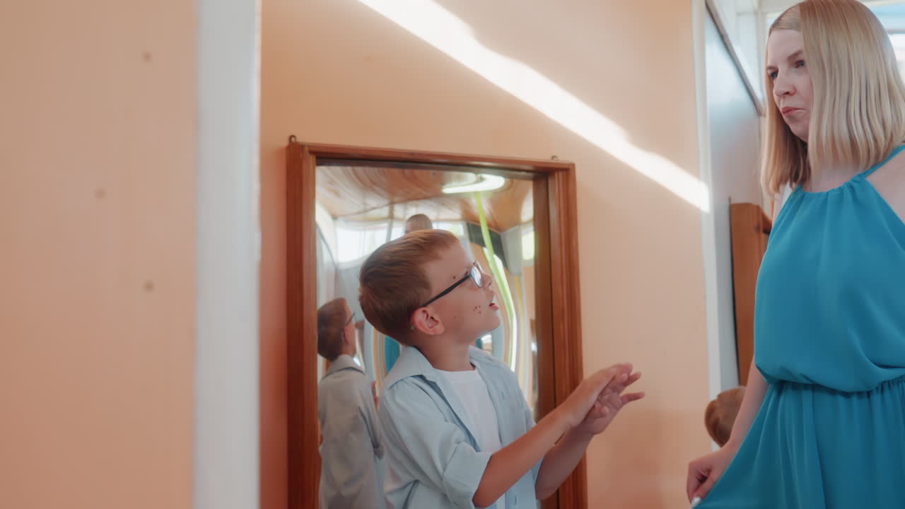 White mother and son family at mirror boy with glasses animatedly explains while mother listens supportively in warm indoor exhibit, playful bonding, storytelling gestures and candid connection